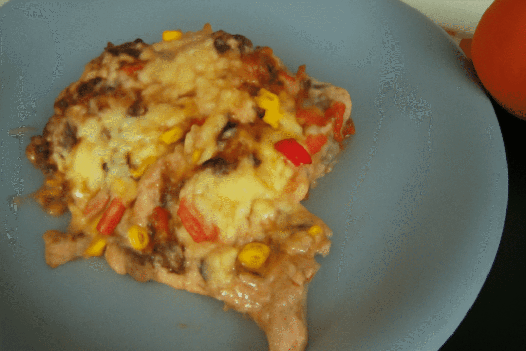 Nahaufnahme eines Nacho-Auflaufs mit geschmolzenem Käse, Mais, roten Paprika und Kidneybohnen auf einem blau-grauen Keramik-Teller.