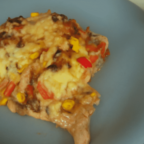 Nahaufnahme eines Nacho-Auflaufs mit geschmolzenem Käse, Mais, roten Paprika und Kidneybohnen auf einem blau-grauen Keramik-Teller.