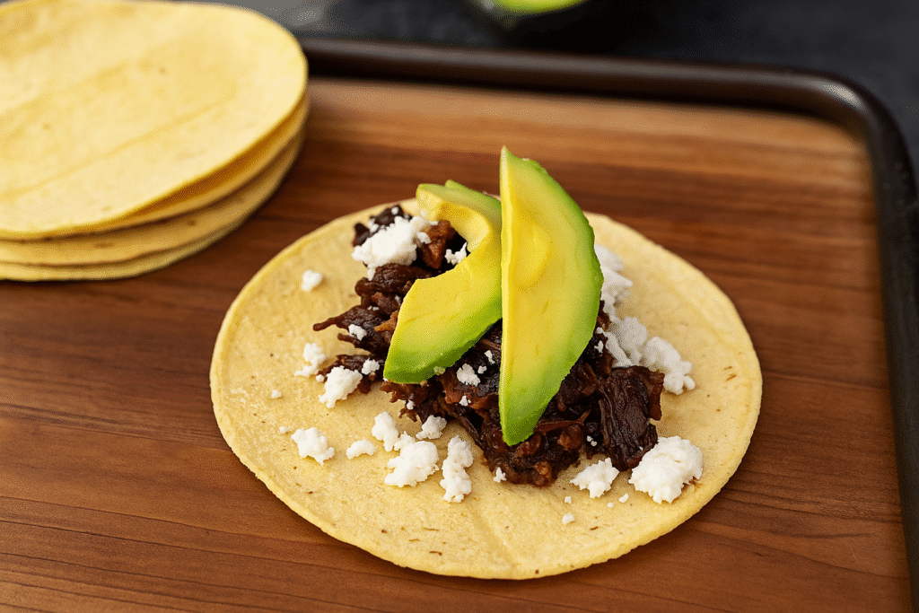 Barbacoa Tacos mit zartem Rindfleisch, frischer Avocado und zerbröseltem Käse auf einer Mais-Tortilla, serviert auf einem Holzbrett.