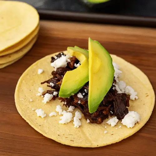 Barbacoa Tacos mit zartem Rindfleisch, frischer Avocado und zerbröseltem Käse auf einer Mais-Tortilla, serviert auf einem Holzbrett.