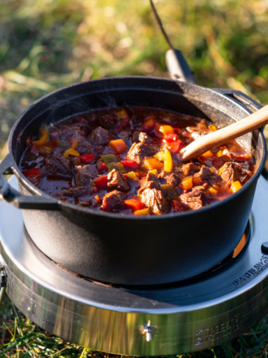 Ein Dutch Oven Gulasch mit zarten Rindfleischstücken, Karotten und Paprika, gekocht in einem Dutch Oven auf einem tragbaren Herd im Freien. Aus dem Topf steigt Dampf auf, was eine warme und einladende Kochszene schafft.
