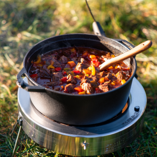 Ein Dutch Oven Gulasch mit zarten Rindfleischstücken, Karotten und Paprika, gekocht in einem Dutch Oven auf einem tragbaren Herd im Freien. Aus dem Topf steigt Dampf auf, was eine warme und einladende Kochszene schafft.