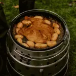 Ein ganzes gewürztes Heuhähnchen liegt auf einem Bett aus Heu im Dutch Oven, umgeben von kleinen Kartoffeln – der Dutch Oven steht auf einem Petromax Atago im Freien.