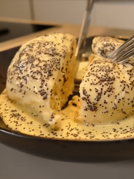 Ein aufgeschnittener Germknödel in einer schwarzen Schale, serviert mit cremiger Vanillesoße und bestreut mit Mohn.