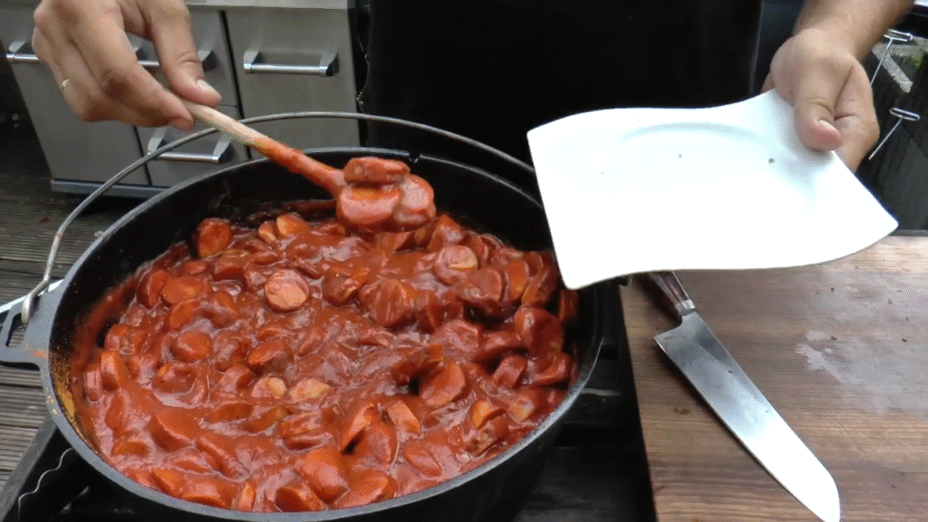 XXL Currywurst topf im Dutch Oven – herzhafter Eintopf mit gegrillten Bratwürsten und würziger Curry-Tomatensoße über offenem Feuer zubereitet.