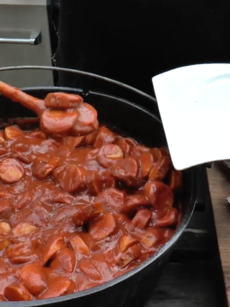 XXL Currywurst topf im Dutch Oven – herzhafter Eintopf mit gegrillten Bratwürsten und würziger Curry-Tomatensoße über offenem Feuer zubereitet.