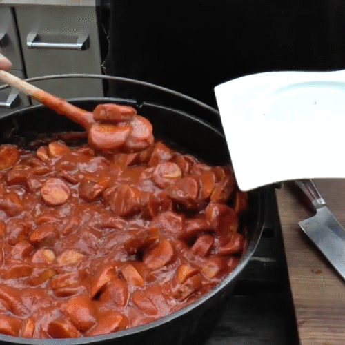 XXL Currywurst topf im Dutch Oven – herzhafter Eintopf mit gegrillten Bratwürsten und würziger Curry-Tomatensoße über offenem Feuer zubereitet.