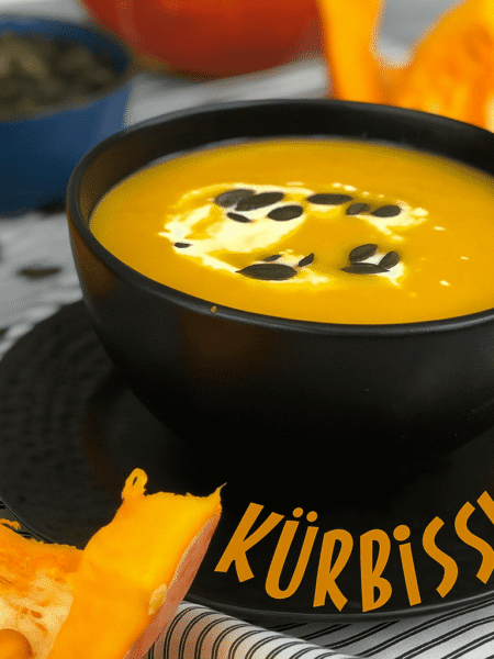 Cremige Kürbissuppe in einer schwarzen Schüssel, garniert mit Kürbiskernen und einem Klecks Sahne, umgeben von frischen Kürbisstücken.