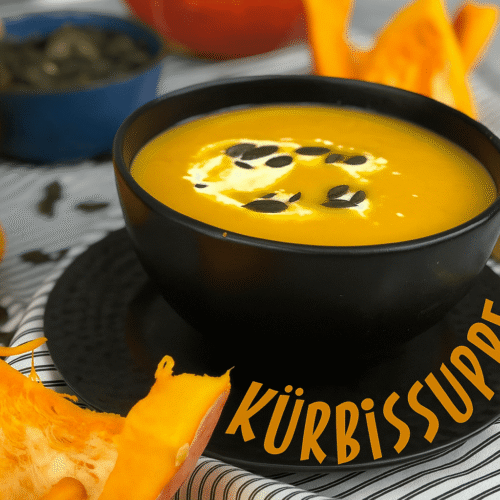Cremige Kürbissuppe in einer schwarzen Schüssel, garniert mit Kürbiskernen und einem Klecks Sahne, umgeben von frischen Kürbisstücken.