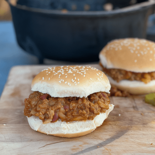 Zwei Sloppy Joe Sandwiches in Sesambrötchen auf einem Holzbrett, mit Hackfleischfüllung und einigen Gurkenscheiben daneben.